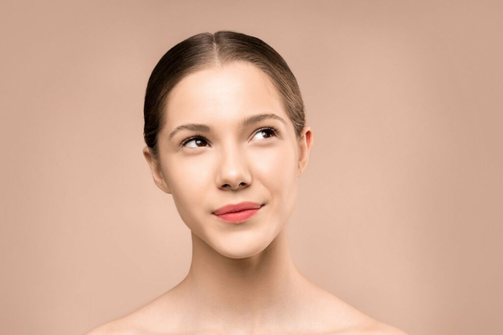 A young woman with radiant skin looks upward against a neutral background, embodying natural beauty.