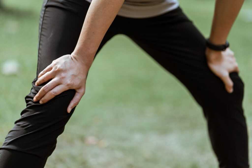 Crop unrecognizable sportsperson standing on recovery posture with hands on knees after active workout in green park